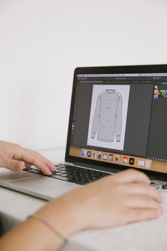 Graphic designer working on clothing design with software on a laptop, focusing on fashion illustration.